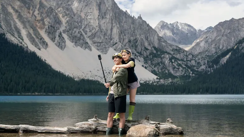 Couple posing on a log in a calm mountain lake while recording with a 360-action camera on a selfie stick Couple posing on a log in a calm mountain lake while recording with a 360-action camera on a selfie stick