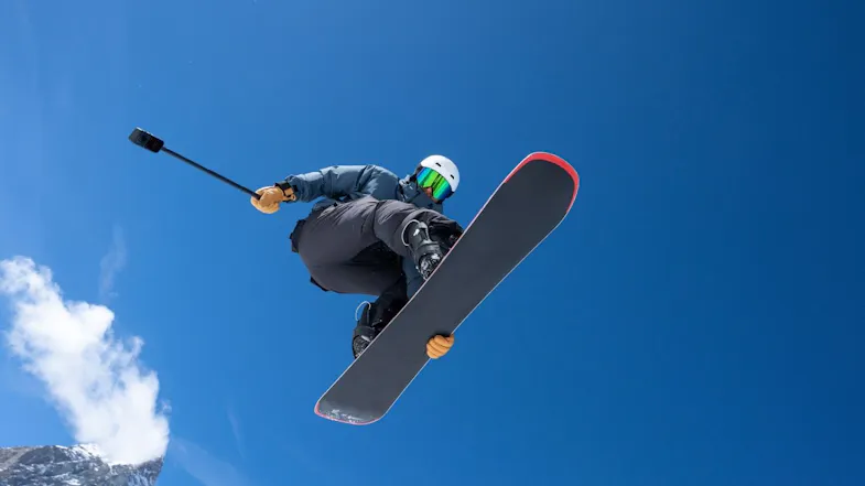 Low-angle view of a snowboarder jumping against a clear blue sky holding an extended action camera selfie stick Low-angle view of a snowboarder jumping against a clear blue sky holding an extended action camera selfie stick