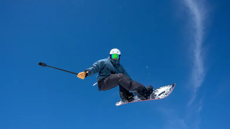 Snowboarder mid-jump against a bright blue sky holding an extended selfie pole with a 360-action camera Snowboarder mid-jump against a bright blue sky holding an extended selfie pole with a 360-action camera