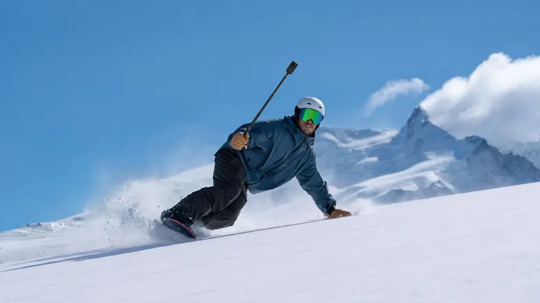 A snowboarder carving down a snowy slope near mountains while holding a 360-action camera on an invisible stick A snowboarder carving down a snowy slope near mountains while holding a 360-action camera on an invisible stick
