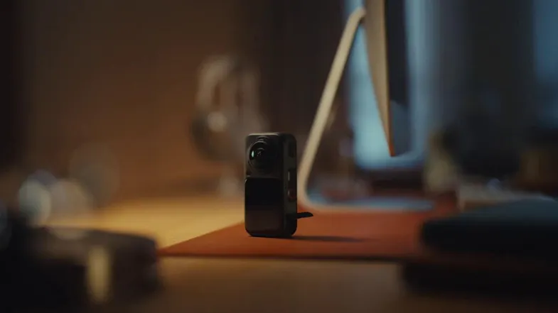 Compact black action camera standing upright on a desk mat next to a desktop monitor in warm indoor light Compact black action camera standing upright on a desk mat next to a desktop monitor in warm indoor light
