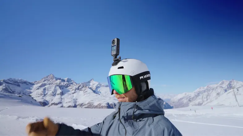 Snowboarder wearing a white helmet with a 360-action camera mounted on top against snowy mountains Snowboarder wearing a white helmet with a 360-action camera mounted on top against snowy mountains