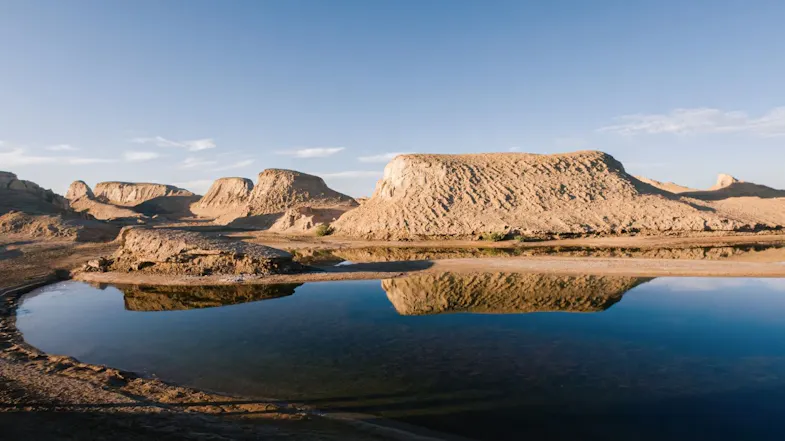 Desert rock formations and sand mounds reflecting a still body of water under a clear blue sky Desert rock formations and sand mounds reflecting a still body of water under a clear blue sky
