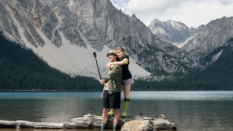 Couple standing on a log in a mountain lake, man holding an extended selfie stick with a 360-action camera Couple standing on a log in a mountain lake, man holding an extended selfie stick with a 360-action camera