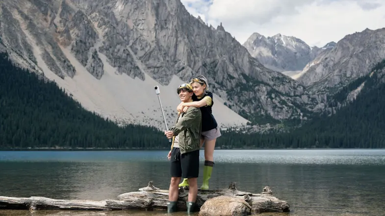 Two people posing on a log in an alpine lake while recording with a 360-action camera on a selfie stick Two people posing on a log in an alpine lake while recording with a 360-action camera on a selfie stick