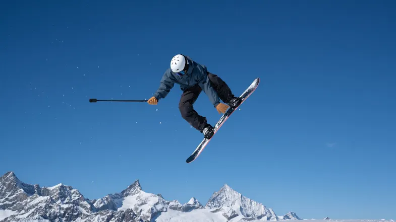 Snowboarder in mid-air above snowy mountains holding an extended selfie stick attached to an action camera Snowboarder in mid-air above snowy mountains holding an extended selfie stick attached to an action camera