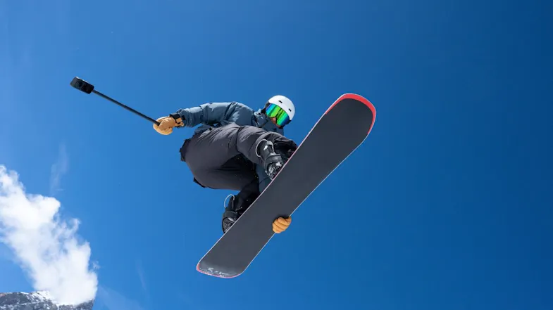 Low-angle view of a snowboarder catching air against a clear blue sky while filming with an action camera Low-angle view of a snowboarder catching air against a clear blue sky while filming with an action camera