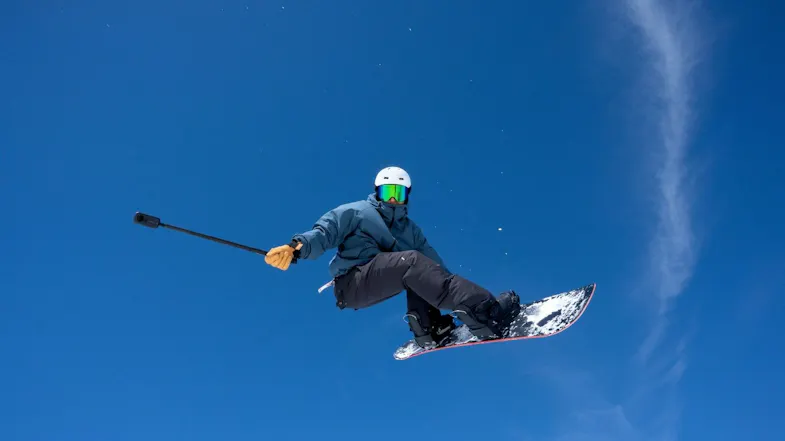 Snowboarder floating through a clear blue sky during a jump while holding a 360 camera on a selfie stick Snowboarder floating through a clear blue sky during a jump while holding a 360 camera on a selfie stick