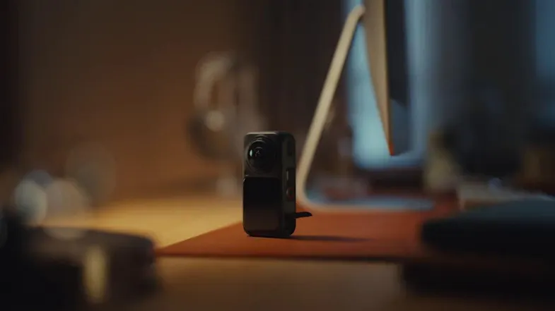 Compact black 360 camera standing upright on a desk mat next to a computer monitor in warm indoor light Compact black 360 camera standing upright on a desk mat next to a computer monitor in warm indoor light