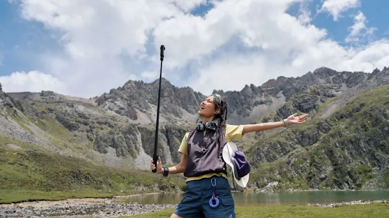Woman standing by a mountain lake holding a long selfie stick with an action camera, outstretched arm Woman standing by a mountain lake holding a long selfie stick with an action camera, outstretched arm