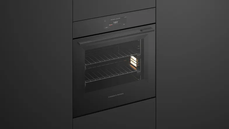 Angled view of a black Fisher & Paykel built-in wall oven with its interior light on, showing wire racks Angled view of a black Fisher & Paykel built-in wall oven with its interior light on, showing wire racks