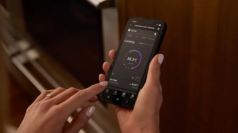 Hands holding a smartphone displaying an oven app with cooking temperature data and a progress circle Hands holding a smartphone displaying an oven app with cooking temperature data and a progress circle