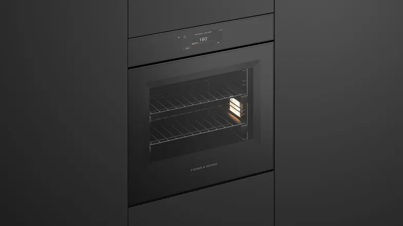 An angled view of a black built-in Fisher & Paykel wall oven with its interior light turned on, showing wire racks An angled view of a black built-in Fisher & Paykel wall oven with its interior light turned on, showing wire racks