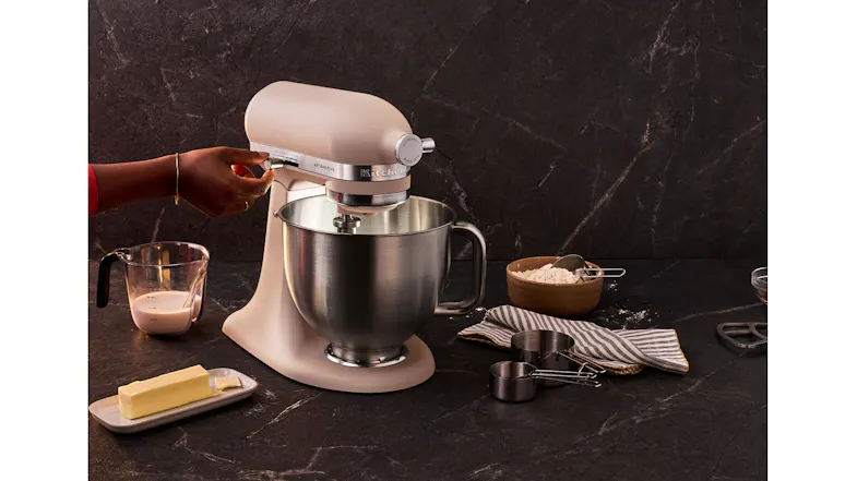 Feathered Pink KitchenAid Artisan Plus Stand Mixer with a stainless steel bowl in a baking preparation setup Feathered Pink KitchenAid Artisan Plus Stand Mixer with a stainless steel bowl in a baking preparation setup