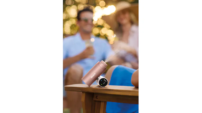 Rose gold Shark ChillPill device resting on a wooden table armrest, spraying a fine mist with blurred people in the background Rose gold Shark ChillPill device resting on a wooden table armrest, spraying a fine mist with blurred people in the background