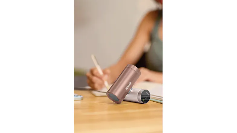 Rose gold Shark ChillPill cooling device angled on a wooden desk with a blurred person writing in the background Rose gold Shark ChillPill cooling device angled on a wooden desk with a blurred person writing in the background