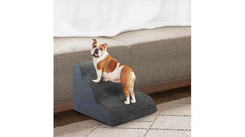 A dog standing on a grey PaWz three-step foam pet stair positioned next to a beige living room sofa A dog standing on a grey PaWz three-step foam pet stair positioned next to a beige living room sofa
