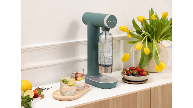 Soda water maker with reusable bottle attached, displayed on a kitchen benchtop beside fresh fruit and flowers Soda water maker with reusable bottle attached, displayed on a kitchen benchtop beside fresh fruit and flowers