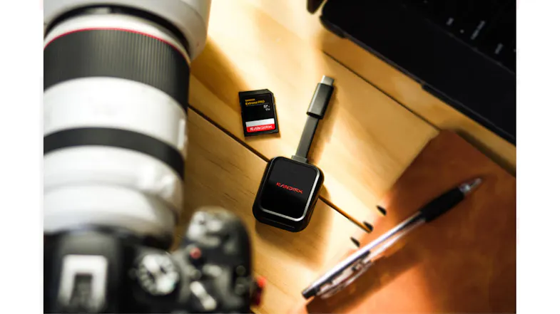 Angled view of a SanDisk Extreme PRO SD Card and a black card reader on a wooden desk beside a camera lens Angled view of a SanDisk Extreme PRO SD Card and a black card reader on a wooden desk beside a camera lens