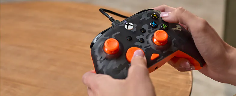 Hands holding a Turtle Beach Rematch wired Xbox controller featuring a black camouflage pattern and bright orange thumbsticks Hands holding a Turtle Beach Rematch wired Xbox controller featuring a black camouflage pattern and bright orange thumbsticks