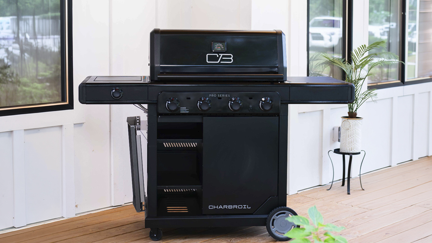 The BBQ is positioned on a wooden deck with the hood closed. The unit features four control knobs and storage
