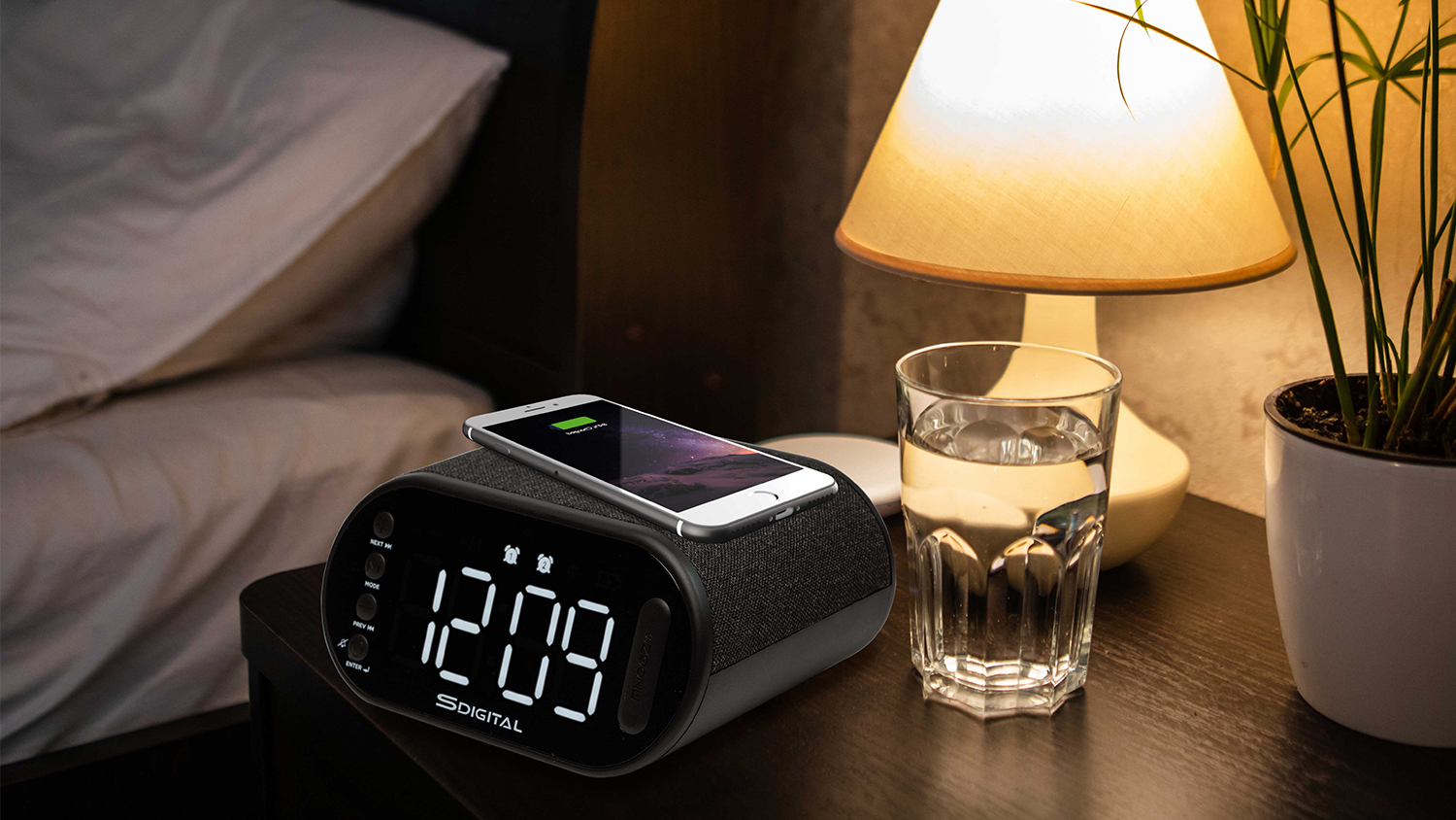 A multifunction clock is on a nightstand with a smartphone above it, showing its wireless charging capability.