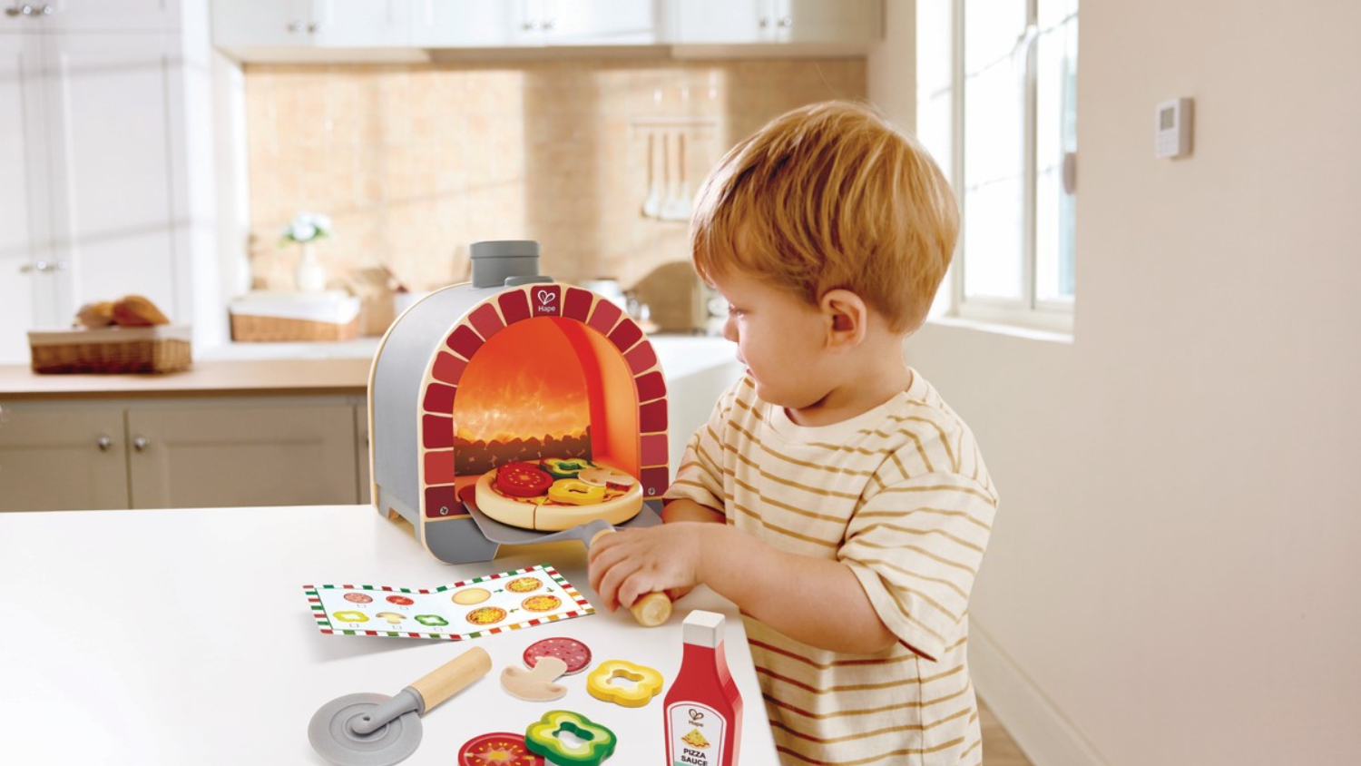 Child playing with toy pizza oven set on kitchen counter with toy ingredients and utensils