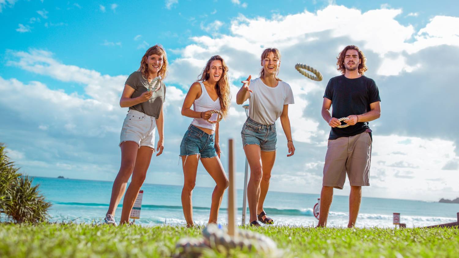 A group of four people playing rope quoits near a beach, one person tossing a ring toward the peg