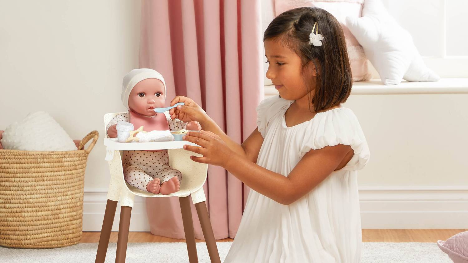 A child feeding a baby doll in a high chair using a blue spoon; other accessories are arranged on the tray