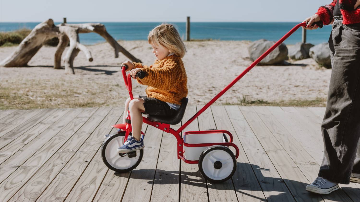 A child being pushed by an adult on the trike along wooden decking at a beachside location