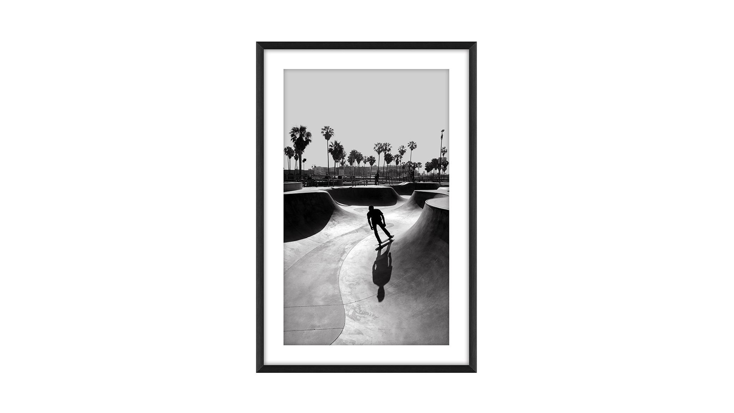 Black and white photo of a skateboarder in motion at a skate park, framed in black, with palm trees and open sky in the background