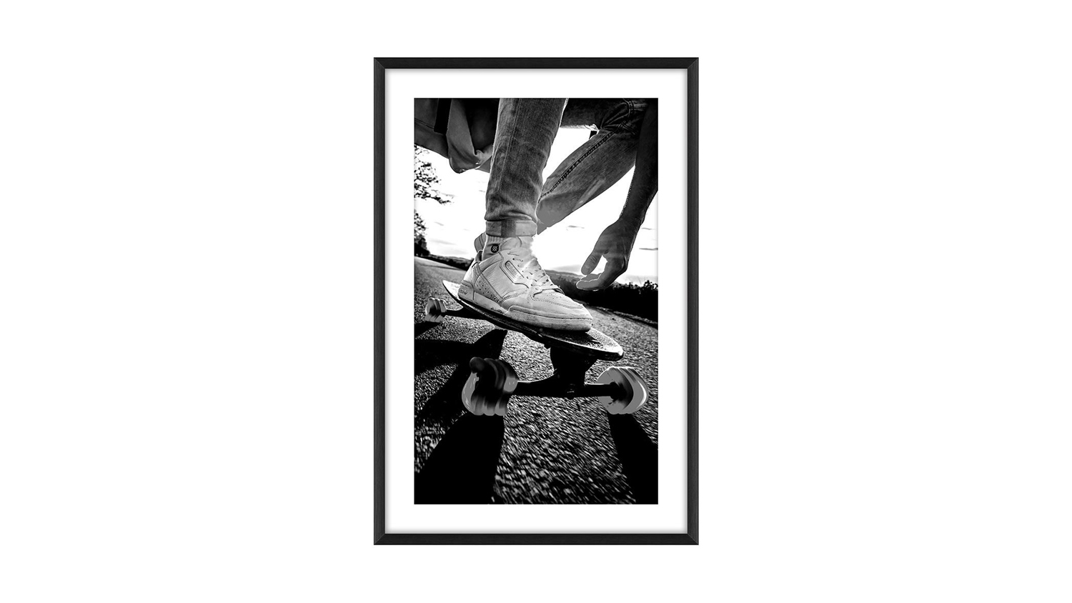 Low-angle black-and-white photo of skateboarder’s feet in sneakers riding on pavement, with shadows, trees, and sky in the background.