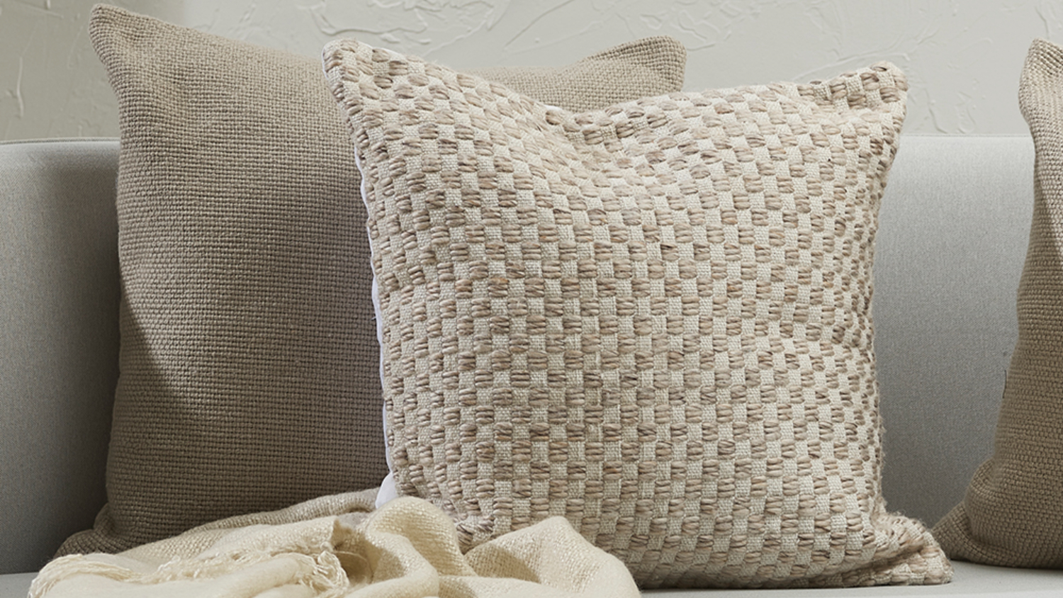 Beige woven cushion on a white sofa next to other neutral coloured cushions.