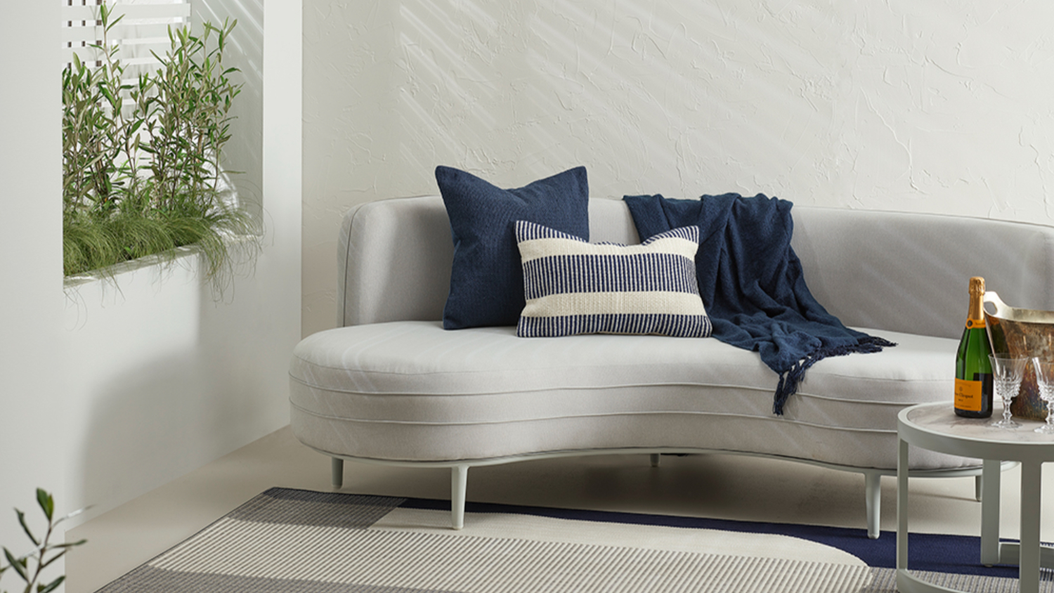 White curved sofa with a striped navy and white cushion and a solid navy cushion, throw blanket, and decorative plants.