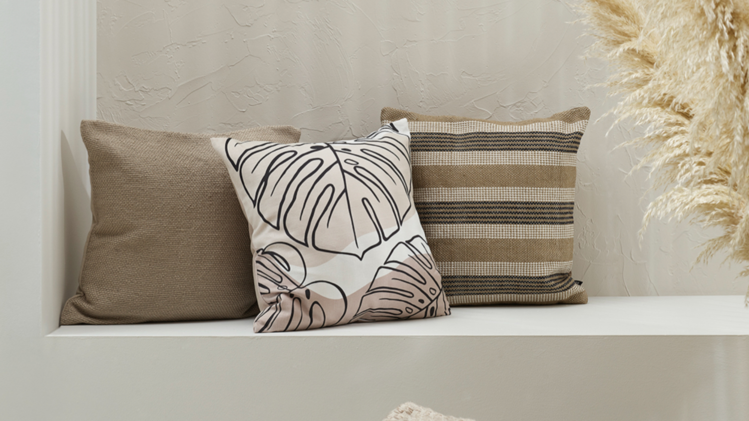 Cushions on a bench, featuring a black and beige leaf-patterned cushion and two textured brown cushions in a neutral-toned setting.