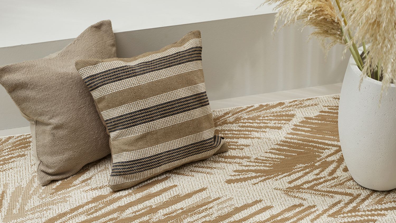 Beige and white rug with leaf pattern, paired with neutral toned cushions on the floor next to pampas grass in vase.