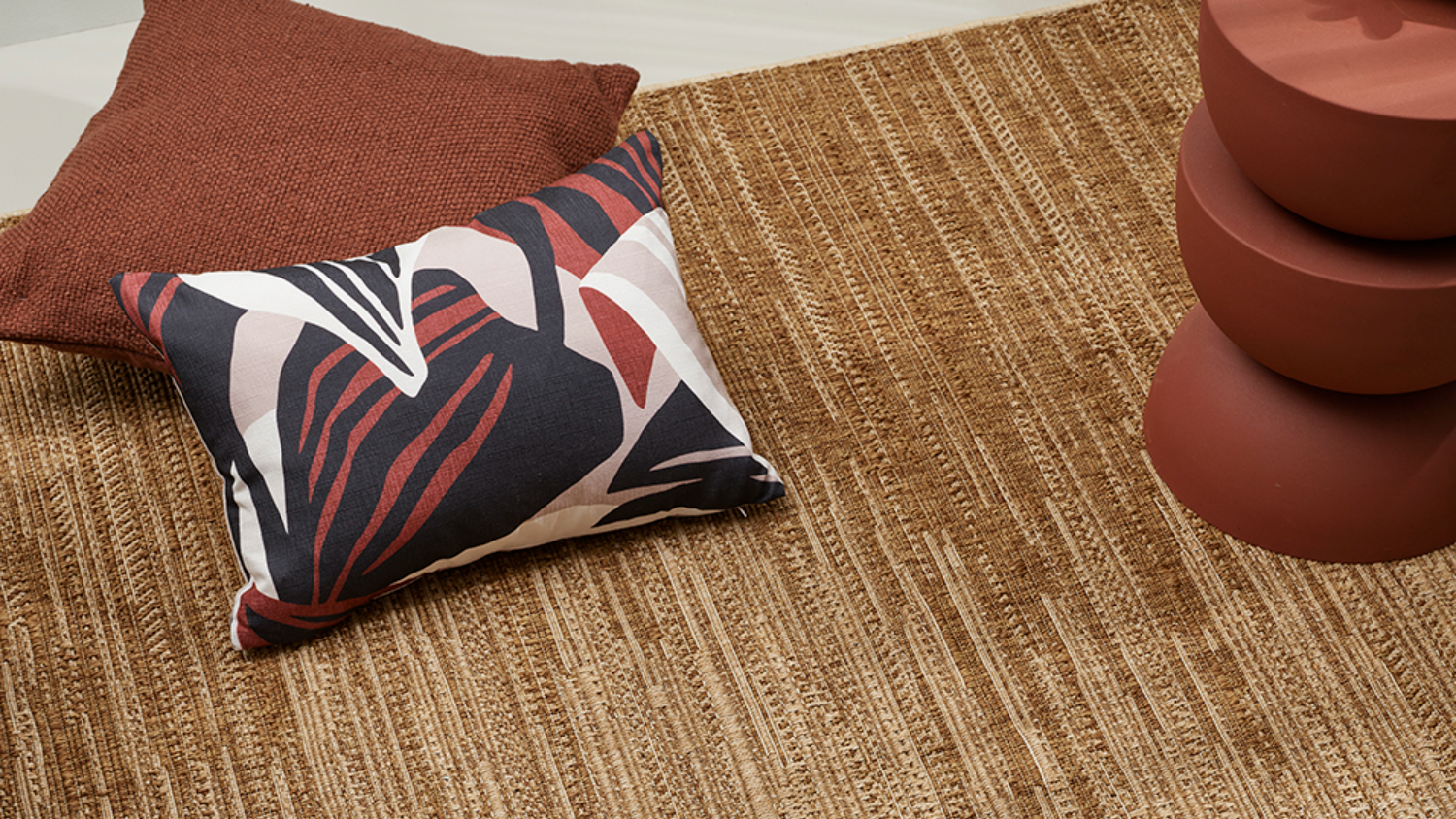Beige woven rug with red pillow, abstract patterned cushion, and stacked round brown decor on the right.