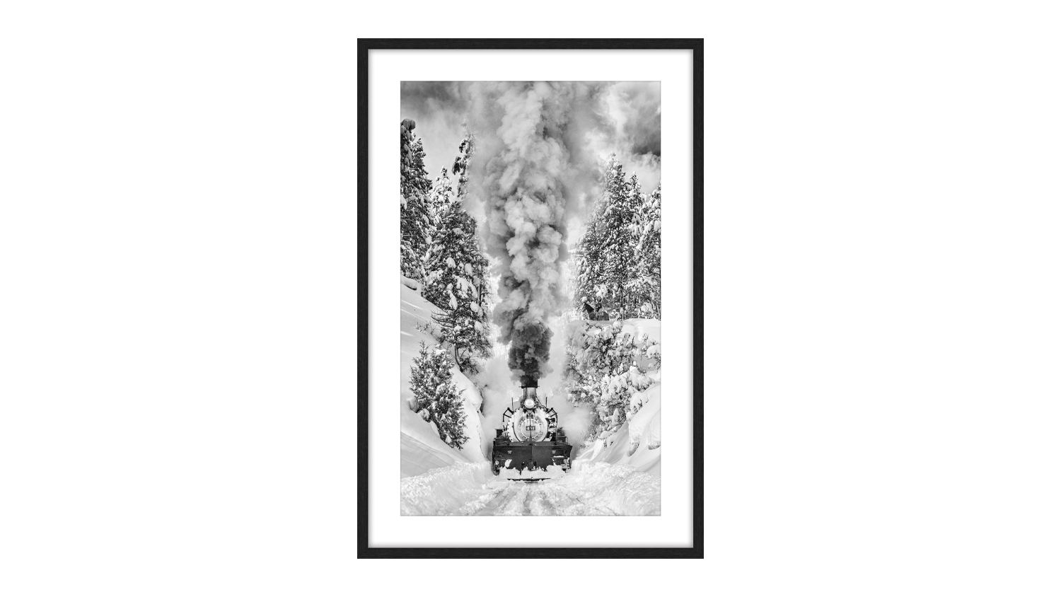 A framed black and white photograph of an old steam locomotive moving through snowy terrain surrounded by trees emitting dense smoke from its chimney.