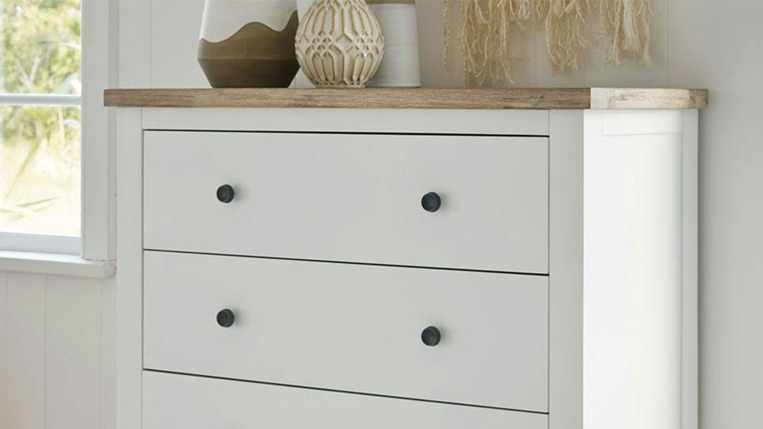 Taylor 4 Drawer Tallboy with décor sitting on top, against a bedroom wall between two windows.