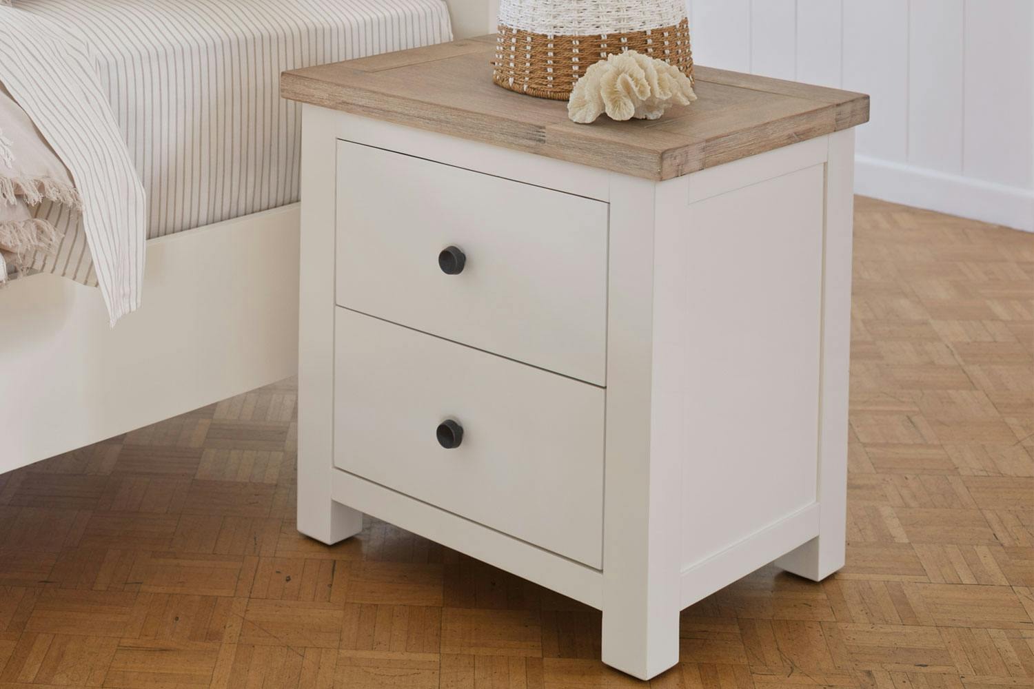 Taylor 2 Drawer Bedside Table shown with a decorative shell and basket sitting on it, beside a bed.