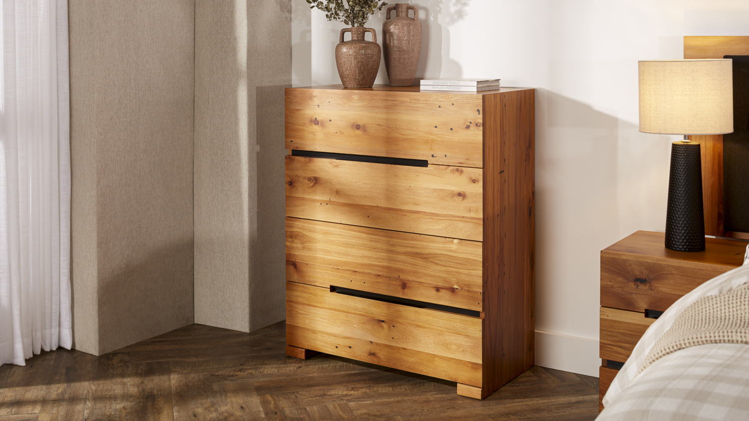 Full view of the rustic wooden four-drawer tallboy next to a bed with a lamp and decorative vases on top