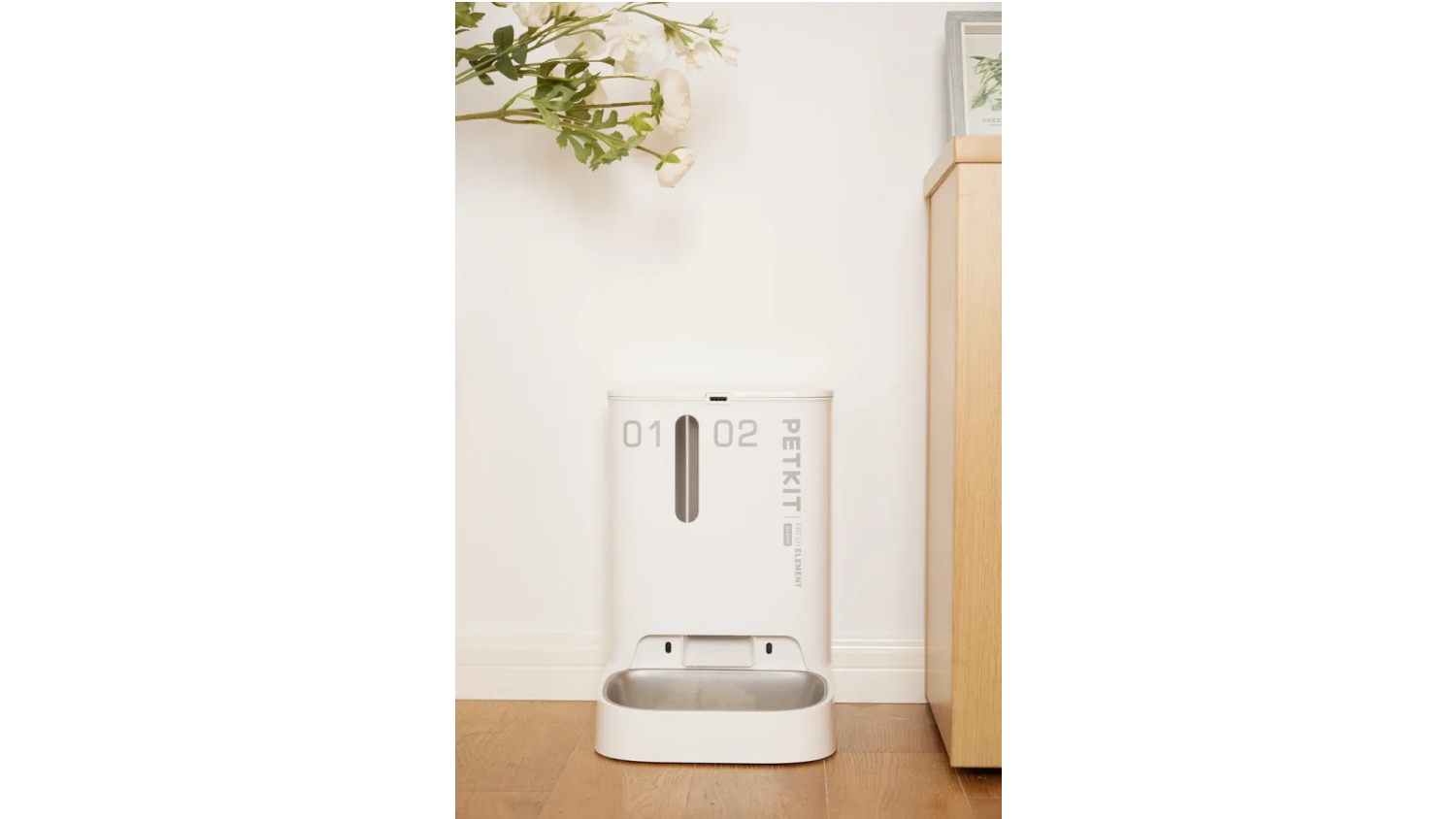 A white PetKit automatic pet feeder is placed on a wooden floor against a white wall
