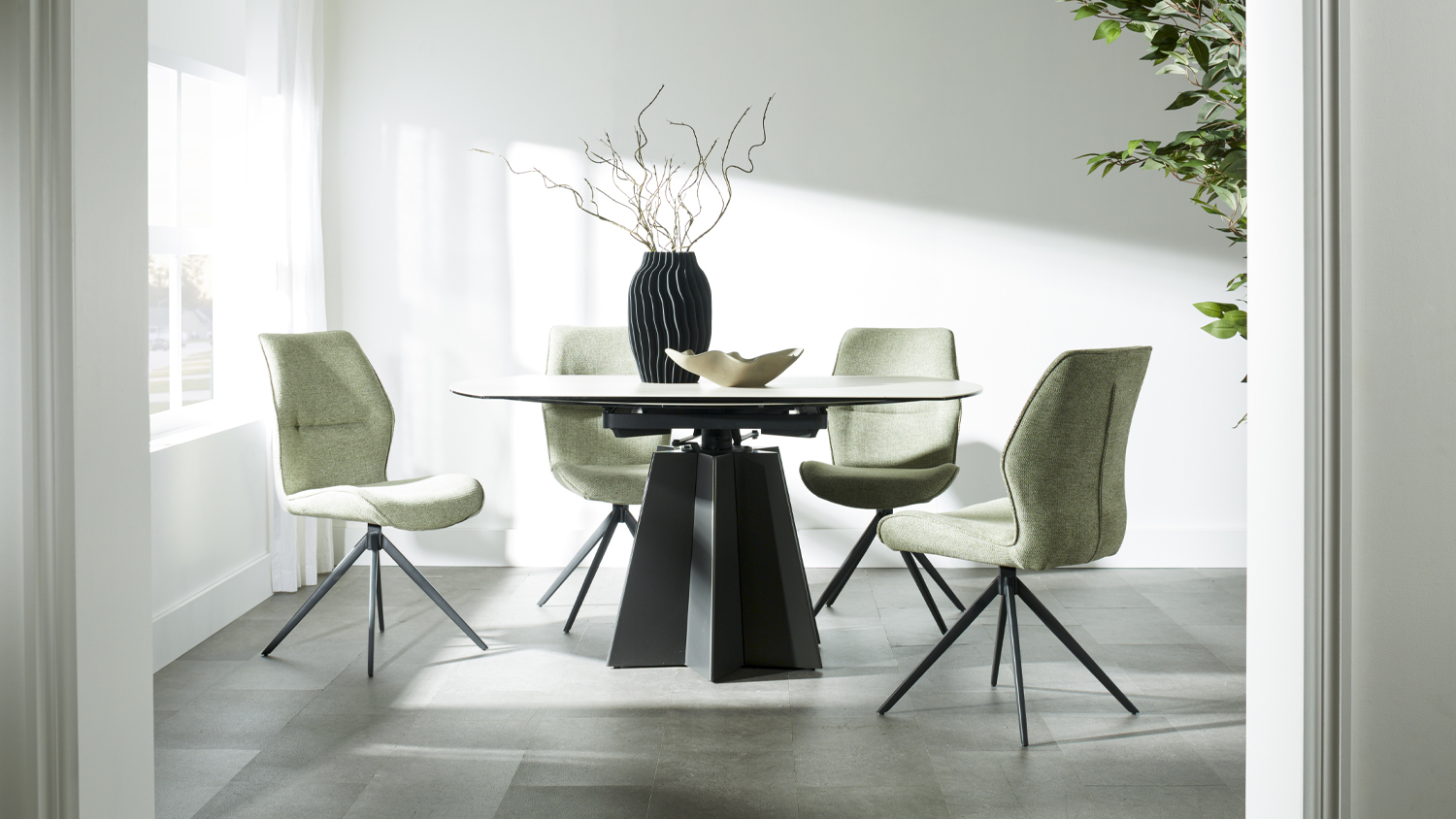 Oval dining table with a light top and black sculptural pedestal base, paired with four olive green fabric chairs