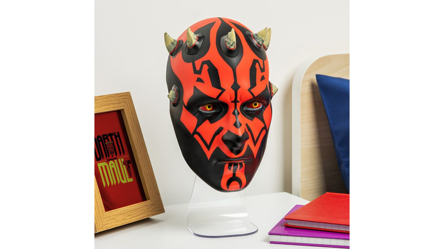 Black and red Paladone novelty wall-mountable mask LED light on a stand, unlit, beside books and a framed print
