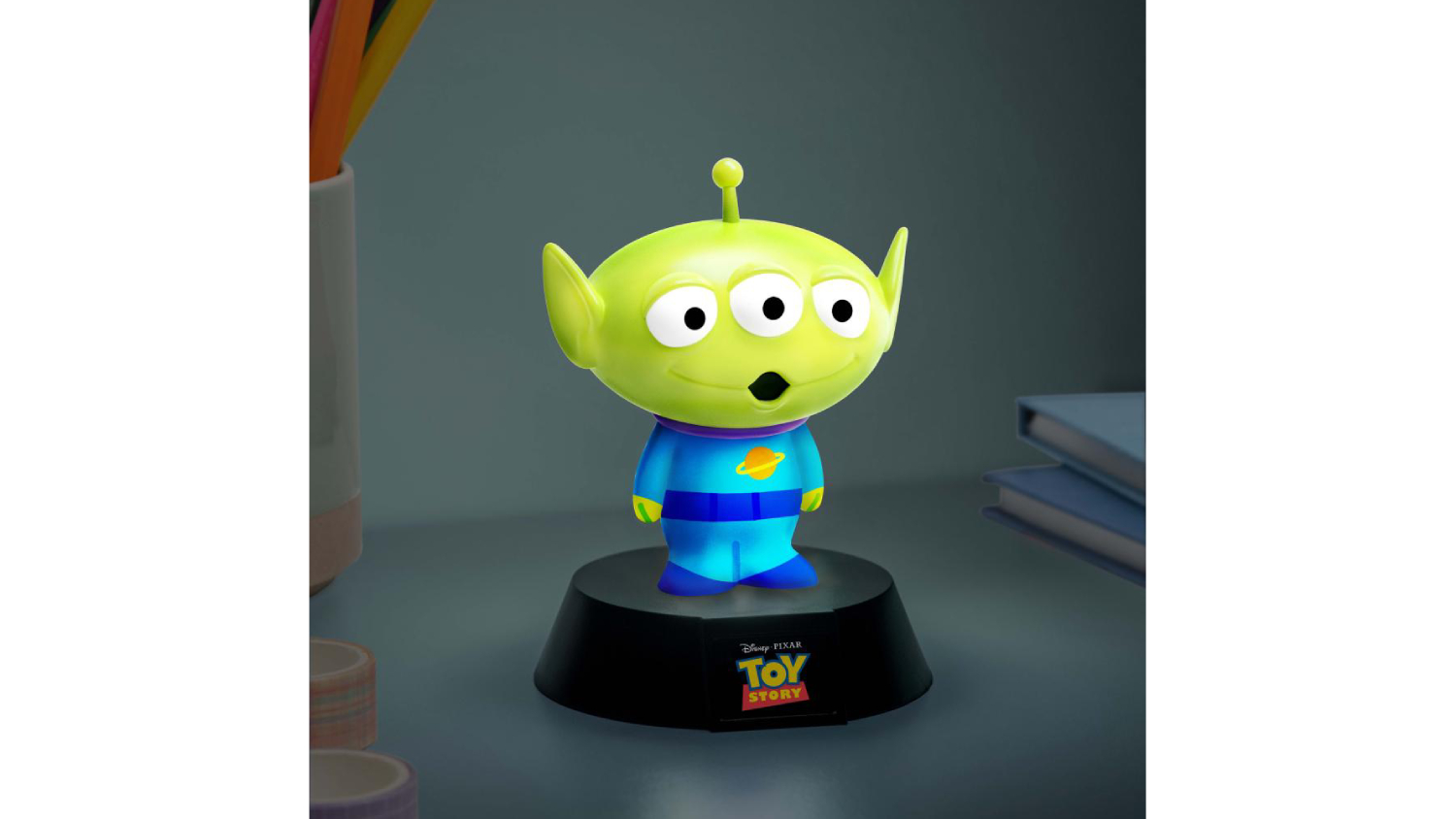 Glowing Toy Story alien Paladone Icon novelty light on a base, with three eyes and an antenna, placed on a desk beside books