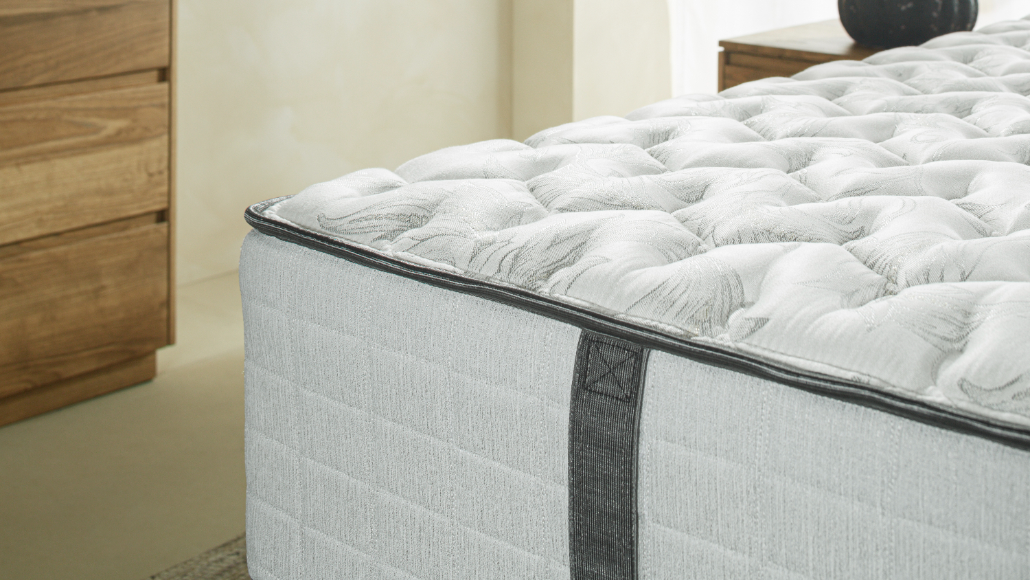 A close-up of a mattress corner showing a tufted white top, grey side panels, and a stitched handle beside a wooden dresser