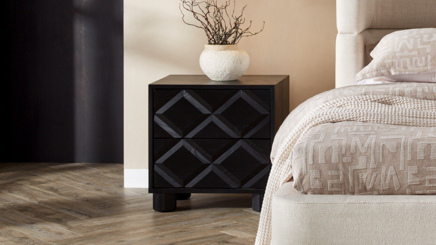 Alexa Diamond 2 Drawer Bedside Table with a vase of branches beside a beige upholstered bed on a wood floor