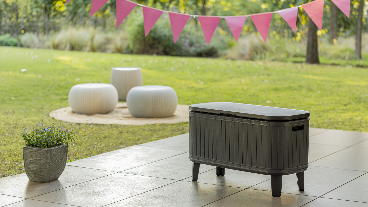 Outdoor patio with a Graphite Keter Bevy Bar Small Storage Cooler with Snack Serving Station, a potted plant, and pink bunting overlooking a grassy garden seating area