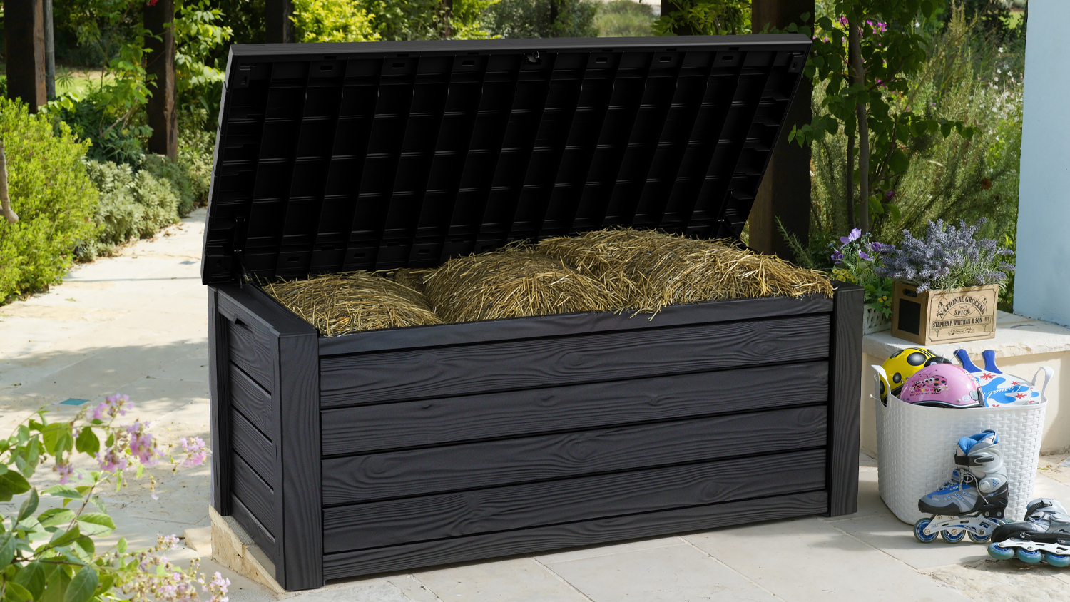 Black Keter Eastwood outdoor storage box with an open lid, filled with straw, on a garden patio surrounded by plants