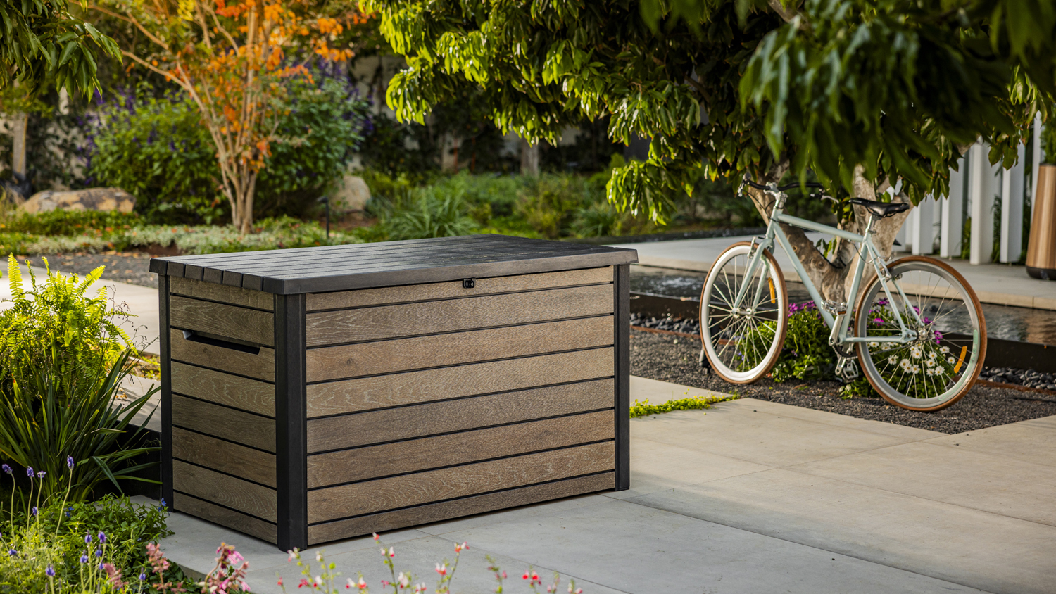 Keter Ontario Large Cushion Storage Box on a patio surrounded by greenery, with a bicycle parked nearby under a tree